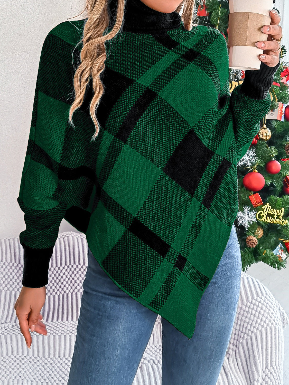 A person adorned in a Trendsi Plaid Turtleneck Batwing Sleeve Sweater, featuring a green and black design crafted from 100% acrylic, holds a coffee cup while standing before a decorated Christmas tree. Exuding a basic style with their long, wavy hair and blue jeans, they achieve an effortlessly chic look. Sweater care instructions suggest machine washing it in cold water.