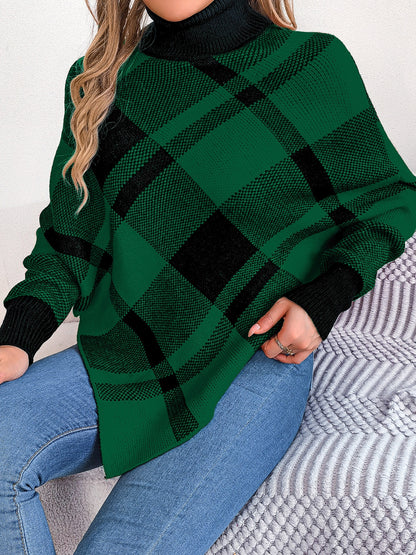 A person wearing a green and black Plaid Turtleneck Batwing Sleeve Sweater by Trendsi, crafted from imported acrylic material, sits on a textured surface. They have long, wavy hair and are paired with blue jeans, with their hand resting on their lap.