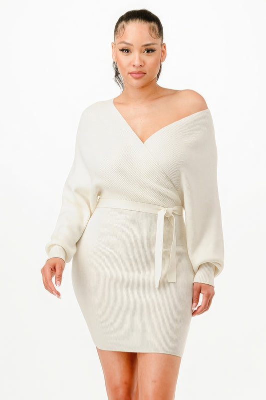 A woman poses against a white background wearing the Keeping It Cozy sweater dress from Serenitylovefashions LLC, which is cream-colored with a ribbed texture and features an off-shoulder design and a tie belt at the waist.