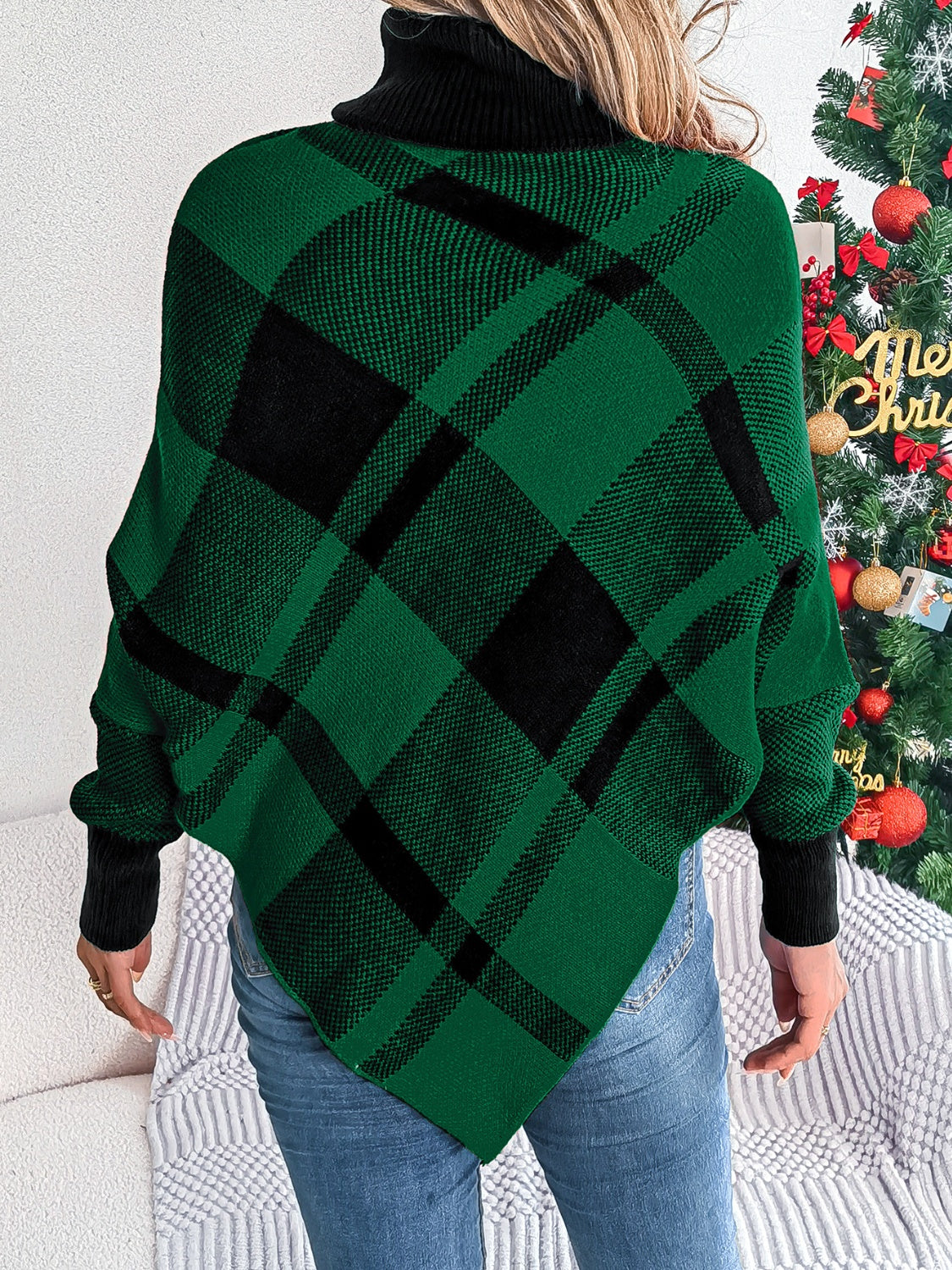 A person stands with their back to the camera, showcasing a Plaid Turtleneck Batwing Sleeve Sweater from Trendsi in green and black hues. They're wearing blue jeans, and a decorated Christmas tree in the background adds holiday cheer to their stylish ensemble.